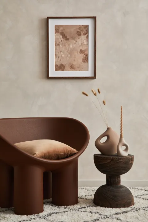 Brown sculptural armchair, carved side table with ceramic vase and framed artwork on a beige wall