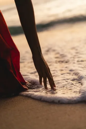 Hand touching sea foam on the sand beside a red dress