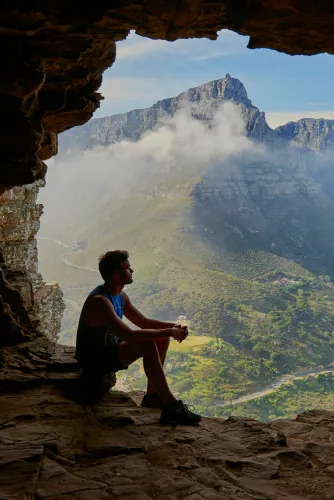 Man sitting in a cave mouth looking out at a mountain landscape
