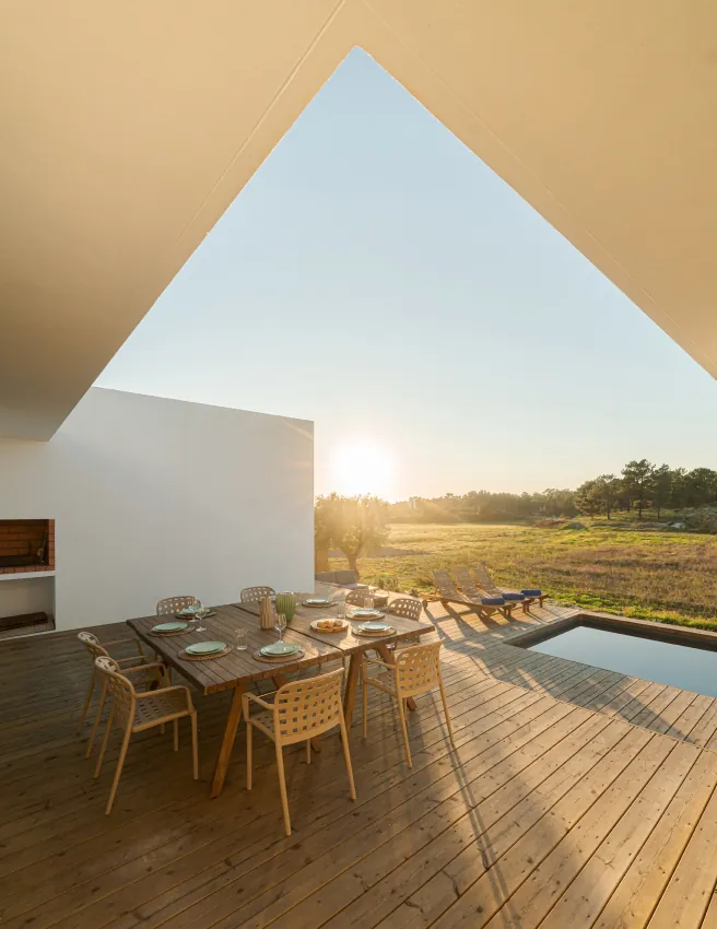 Dining table on the timber terrace of a modern white villa beside a pool at sunset