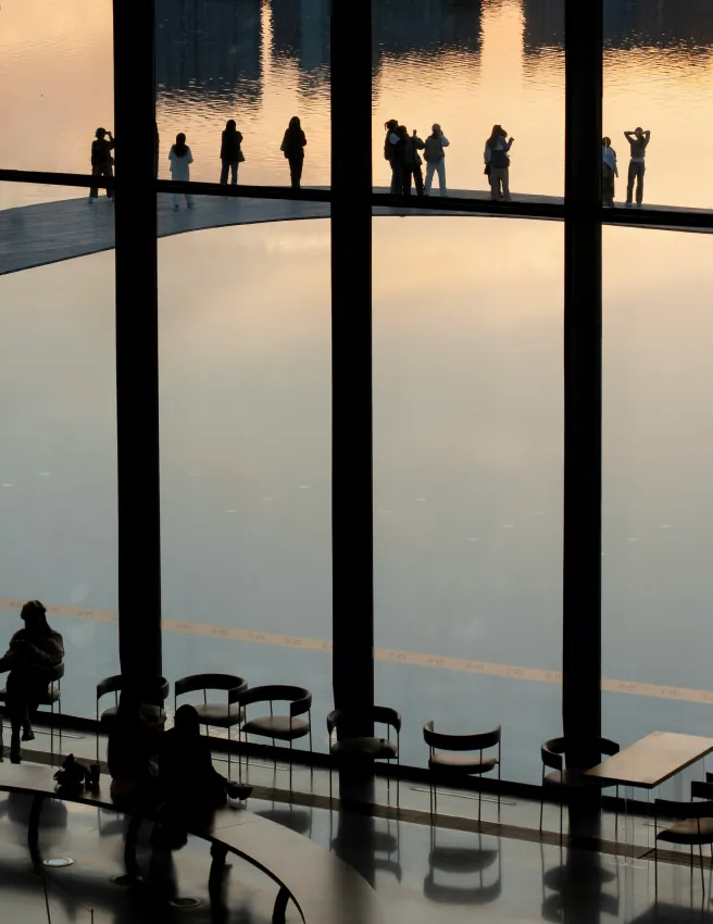 Silhouetted people on a walkway and in a café behind tall windows overlooking water at sunset