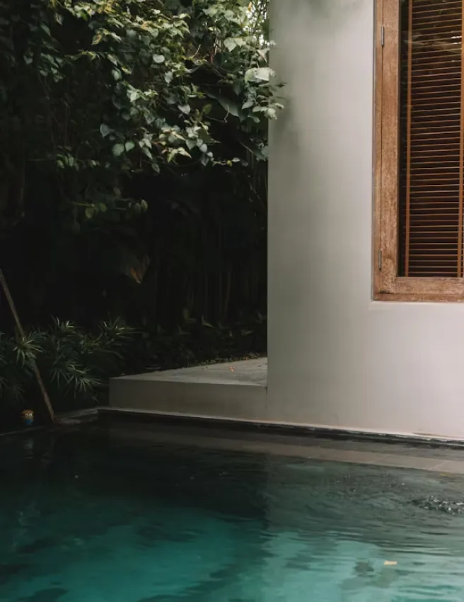 Turquoise pool beside a white rendered wall with a wooden shutter and greenery