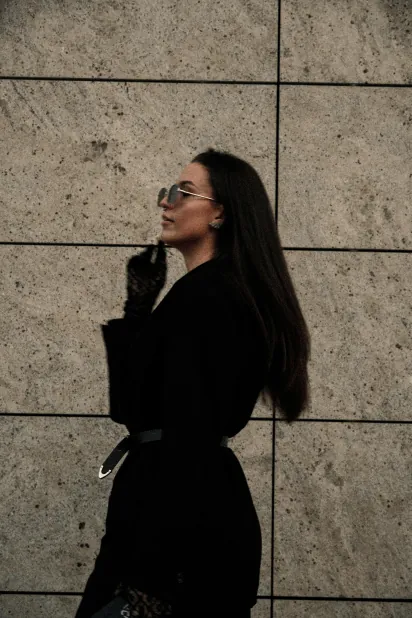 Woman with long dark hair and sunglasses in a black coat against a stone wall
